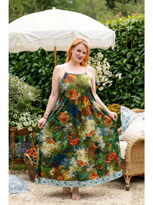 A woman stands barefoot outdoors in a garden setting under a large white patio umbrella, wearing a sleeveless maxi dress with a dense floral print in green, orange, cream, and blue. A wicker chair with pillows is visible to the right. | Bliss by Cori