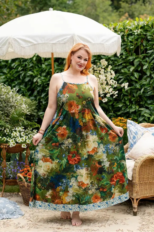 A woman stands barefoot outdoors in a garden setting under a large white patio umbrella, wearing a sleeveless maxi dress with a dense floral print in green, orange, cream, and blue. A wicker chair with pillows is visible to the right. | Bliss by Cori