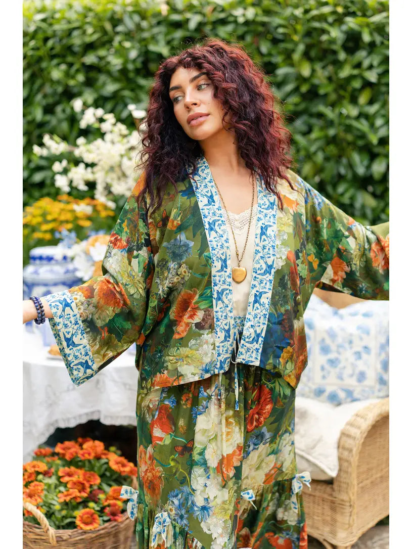 A woman wearing a bohemian-style floral print kaftan robe and matching skirt in a garden setting. The outfit features a green base with orange, blue, and yellow floral patterns, a blue patterned trim | Bliss by Cori