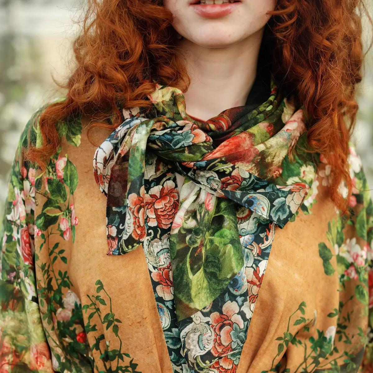 A close-up of a person with curly red hair wearing a tan top and a floral print scarf tied around the neck. The scarf features green leaves and red, pink, and white flowers on a dark background | Bliss by Cori