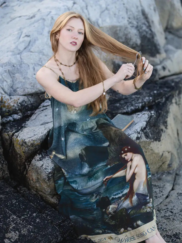 Seated model arranging the flowing skirt of the Siren mermaid maxi dress on shoreline rocks | Bliss by Cori