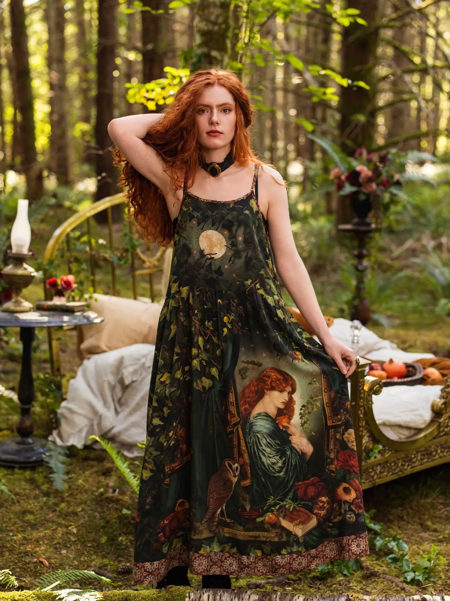 Full-length front view of red-haired model standing in mossy forest wearing Solstice Bohéme Maxi Slip Dress, showing full Rossetti Proserpine portrait print with moon and ivy, antique brass bed and oil lamp behind — Bliss by Cori
