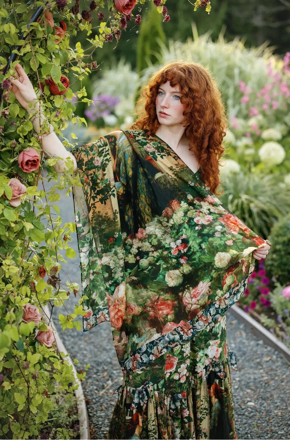 A woman with long red hair stands in a garden wearing a flowing floral dress with wide draped sleeves. The dress features a dense botanical print in green, orange, pink, and cream tones | Bliss by Cori