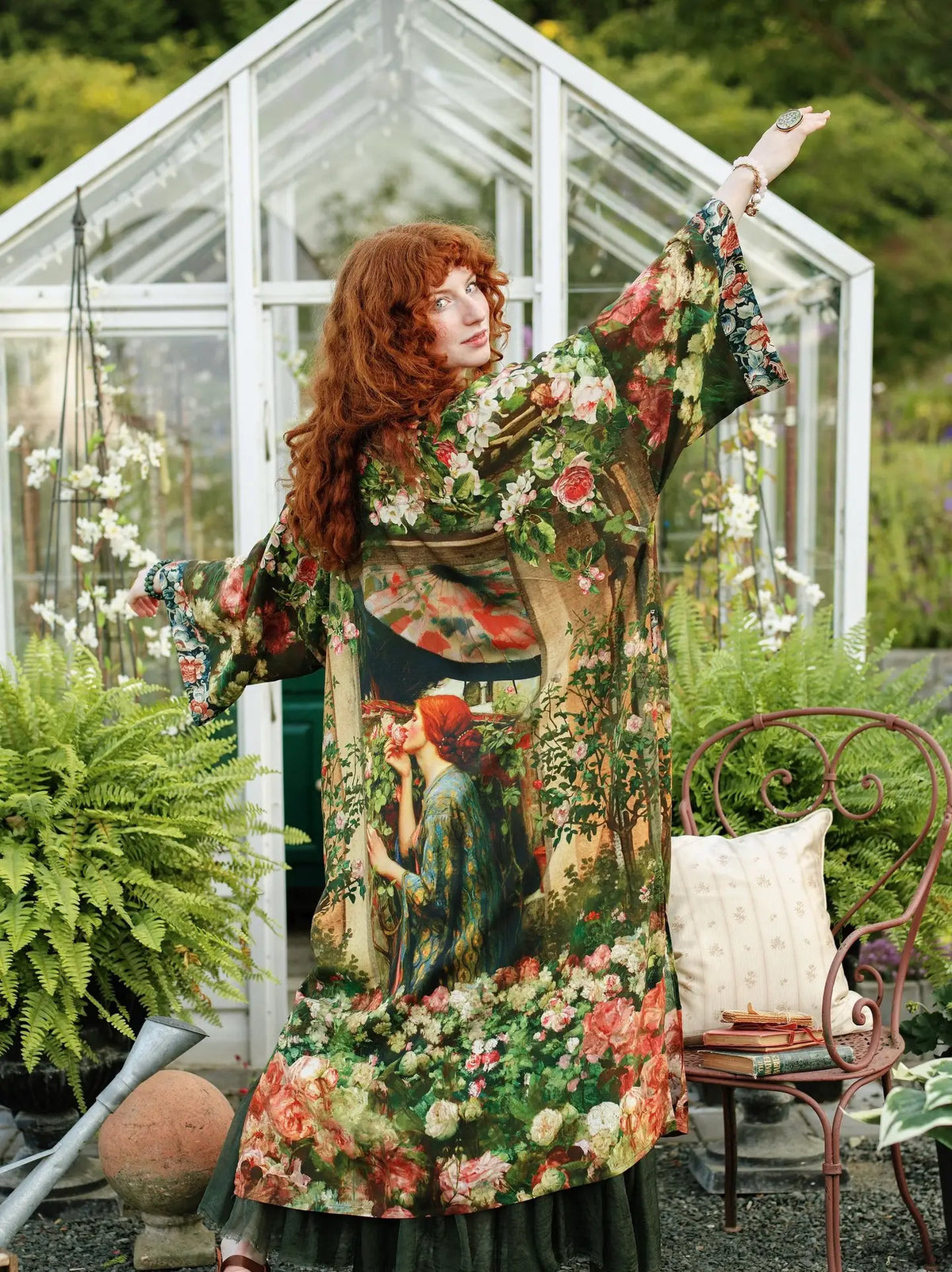 A woman with long red hair stands in a greenhouse garden wearing a long-sleeved, wide-neck kaftan dress with a detailed botanical and figure print. She has one arm raised in a pose, with a rustic metal chair and green plants in the background | Bliss by Cori