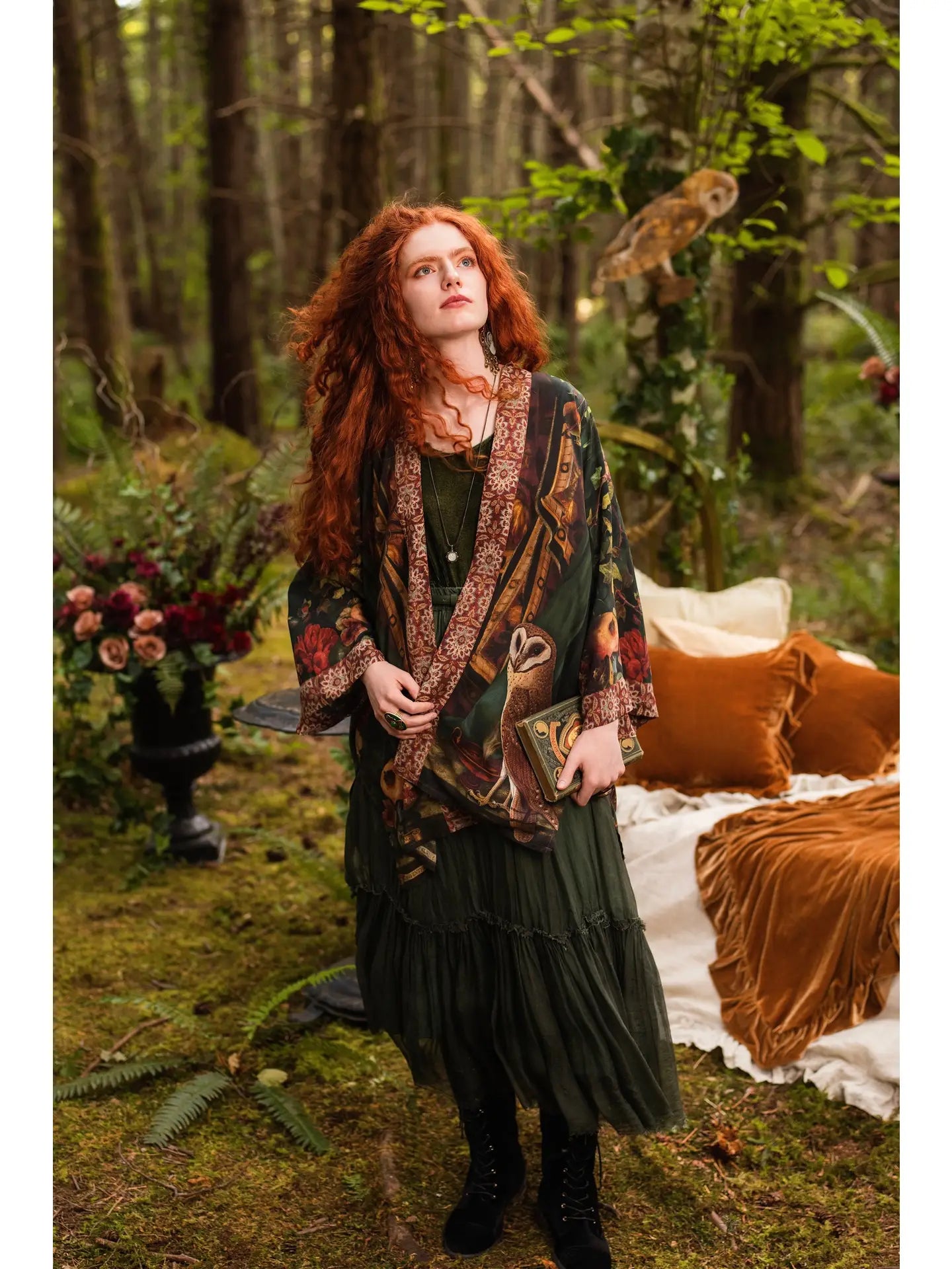 Red-haired model in enchanted forest wearing Solstice Matinée Mid-Length Duster Kimono open over black dress, holding pomegranate, barn owl perched in trees behind, velvet cushions and candles nearby — Bliss by Cori
