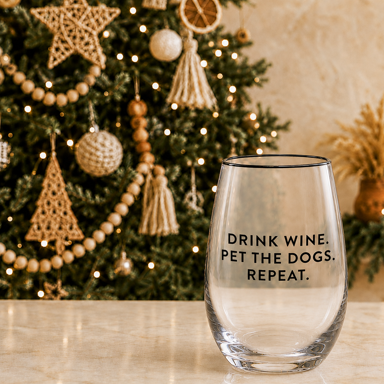 Clear stemless wine glass with black rim and Drink Wine Pet the Dogs Repeat black text on marble counter in front of a boho Christmas tree with wood bead garland macrame tassels and warm fairy lights – Bliss by Cori
