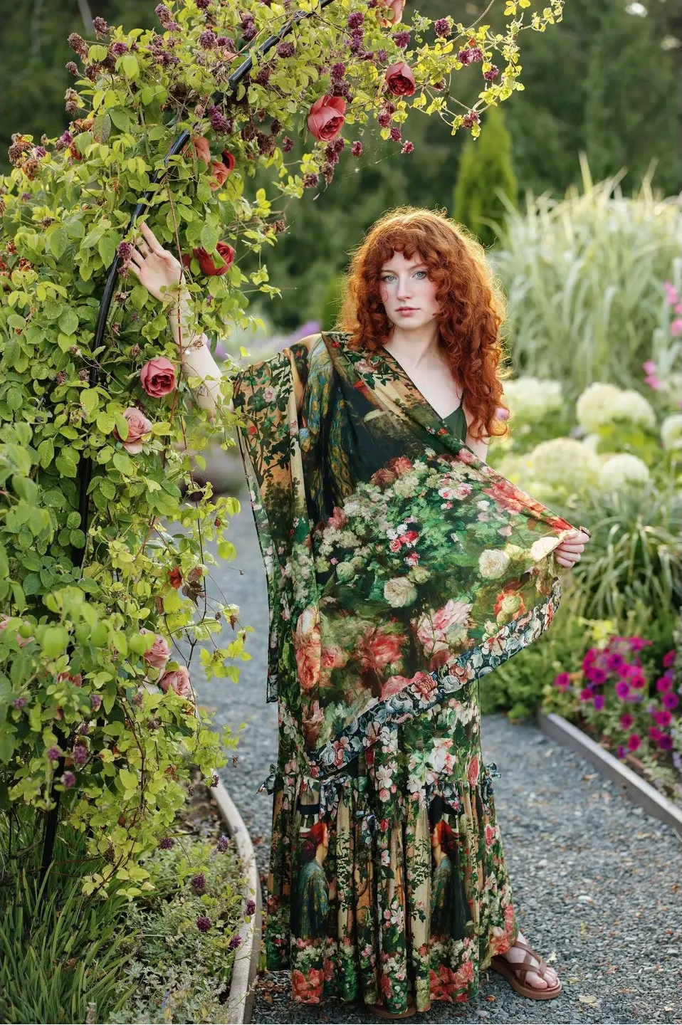 A woman with long curly red hair stands in a garden wearing a long, flowing floral dress with wide draped sleeves. She is posed beside a flowering vine arch in a landscaped garden path | Bliss by Cori