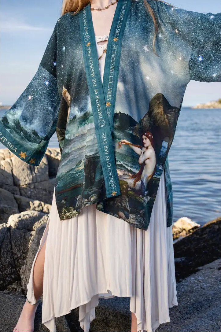 A woman standing outdoors by the water wears a loose, open-front kimono-style cardigan with a celestial and mythological print | Bliss by Cori
