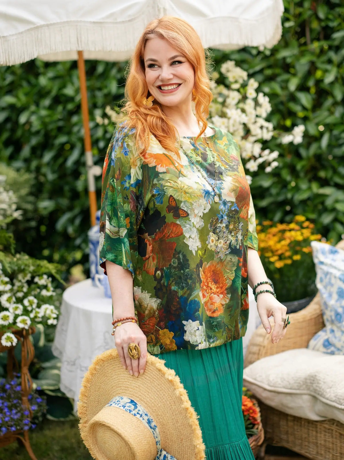Red-haired woman holding a straw hat while wearing a green floral-and-bird print Luxe Tee tunic over a teal skirt in a garden setting | Bliss by Cori