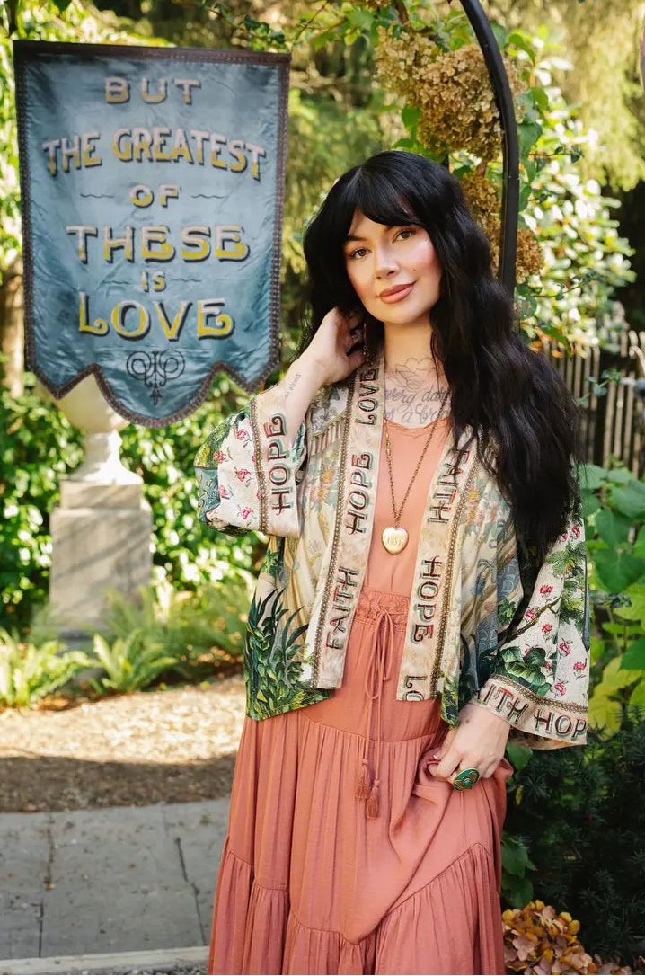 Model wearing Book of Love cropped kimono over a coral dress beside a blue love banner in a garden | Bliss by Cori