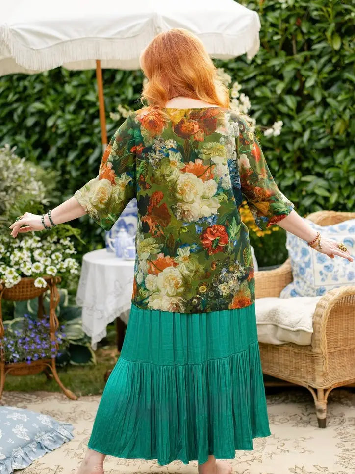 A woman with red hair stands outdoors in a garden setting, viewed from behind, wearing a floral long-sleeve top and a pleated green skirt. The background features a white patio umbrella, a wicker chair, and lush green foliage | Bliss by Cori
