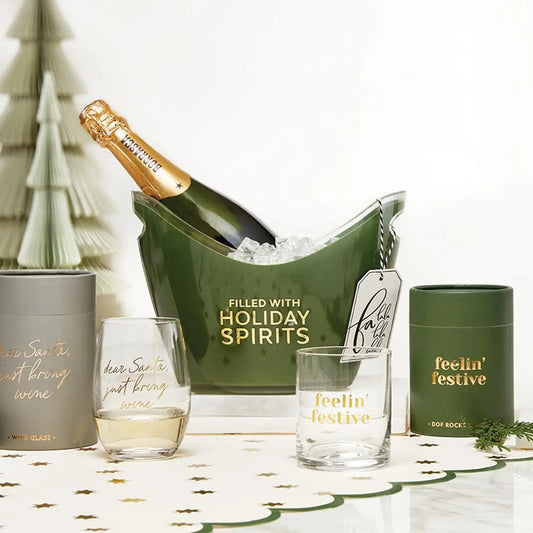 Dear Santa Just Bring Wine stemless wine glass with gold foil script styled beside a Filled with Holiday Spirits champagne bucket Feelin Festive rocks glass and gray gift tube with paper Christmas trees – Bliss by Cori
