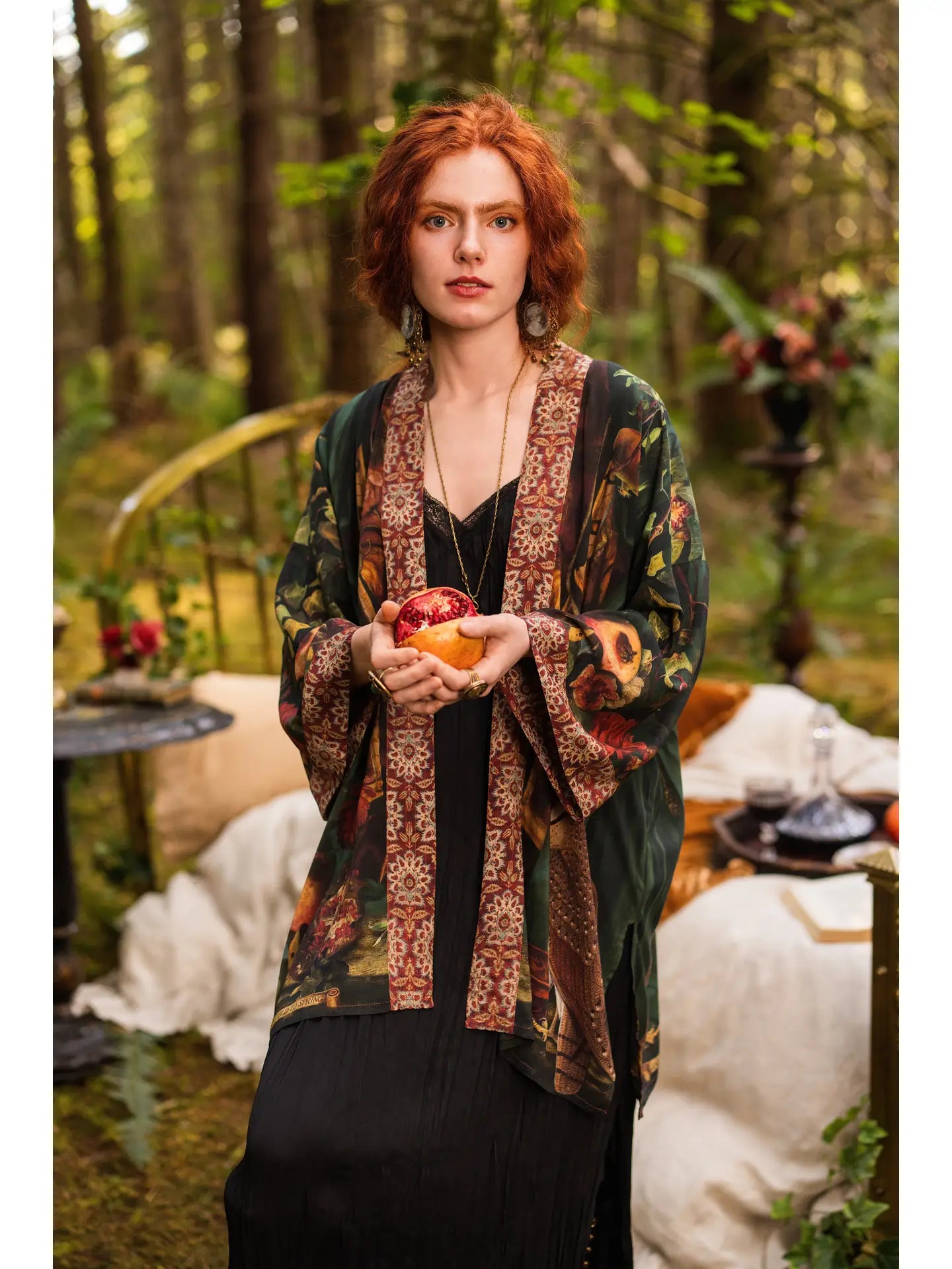 Front view of short red-haired model in forest wearing Solstice Matinée Mid-Length Duster Kimono open over black dress, holding pomegranate, tapestry border trim visible, enchanted outdoor bedroom with velvet cushions behind — Bliss by Cor
