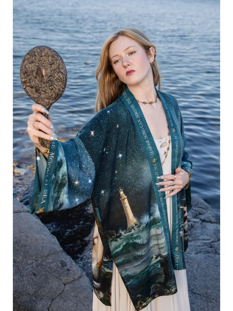 A woman stands on rocks by the water wearing a dark teal kimono-style cardigan with a celestial and lighthouse print. She holds an ornate round hand mirror | Bliss by Cori