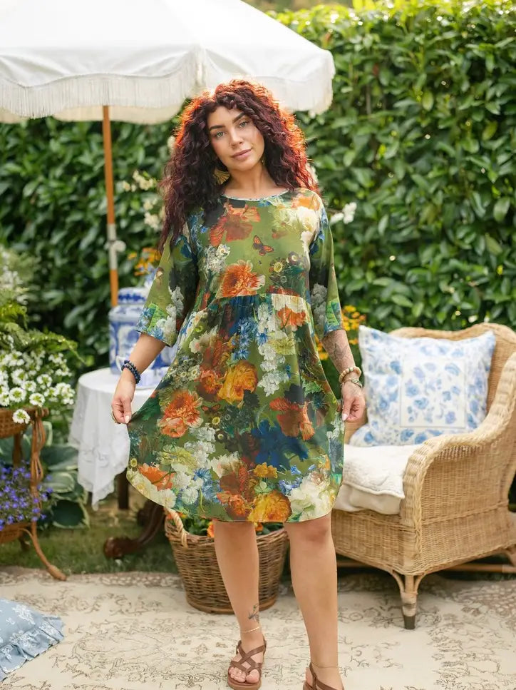 Red-haired woman wearing a green floral artist dress tunic and holding a blue-and-white vase of white flowers in a garden setting | Bliss by Cori