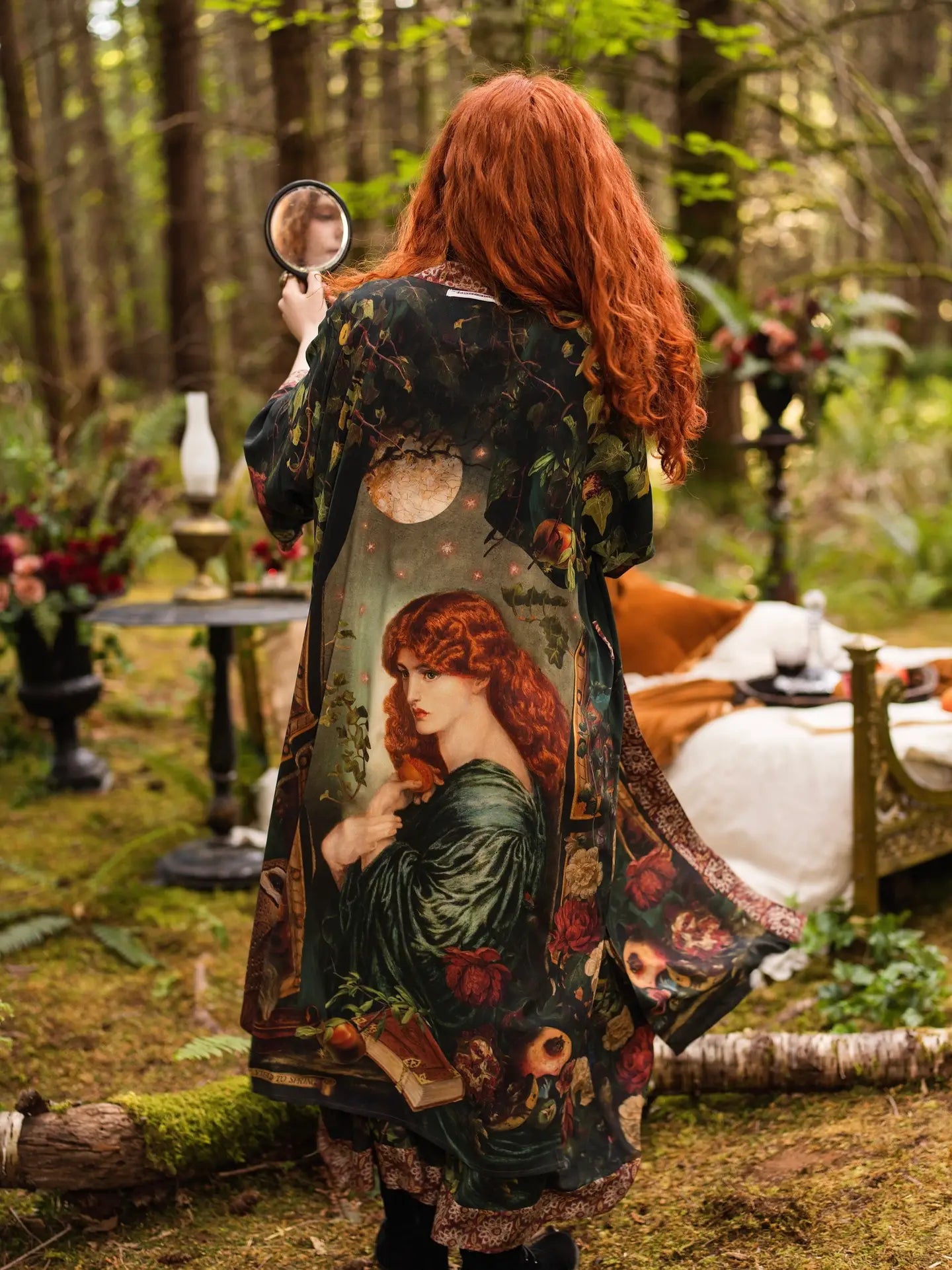 Back view of model holding hand mirror in forest glade wearing Solstice Opera Duster Kimono, full Rossetti Proserpine and moon print visible, enchanted outdoor bedroom with oil lamps behind — Bliss by Cori