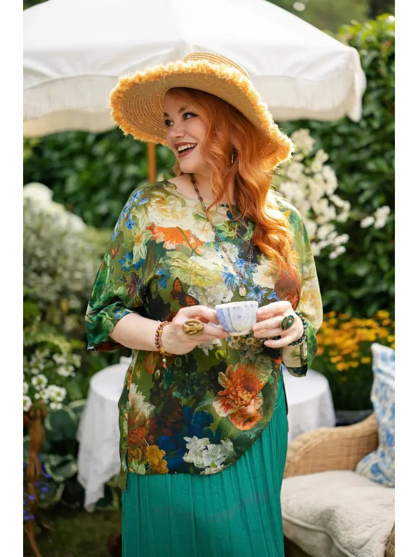 Red-haired woman in a wide-brimmed straw hat holding a teacup, wearing a green floral-and-bird print Luxe Tee tunic over a teal skirt | Bliss by Cori