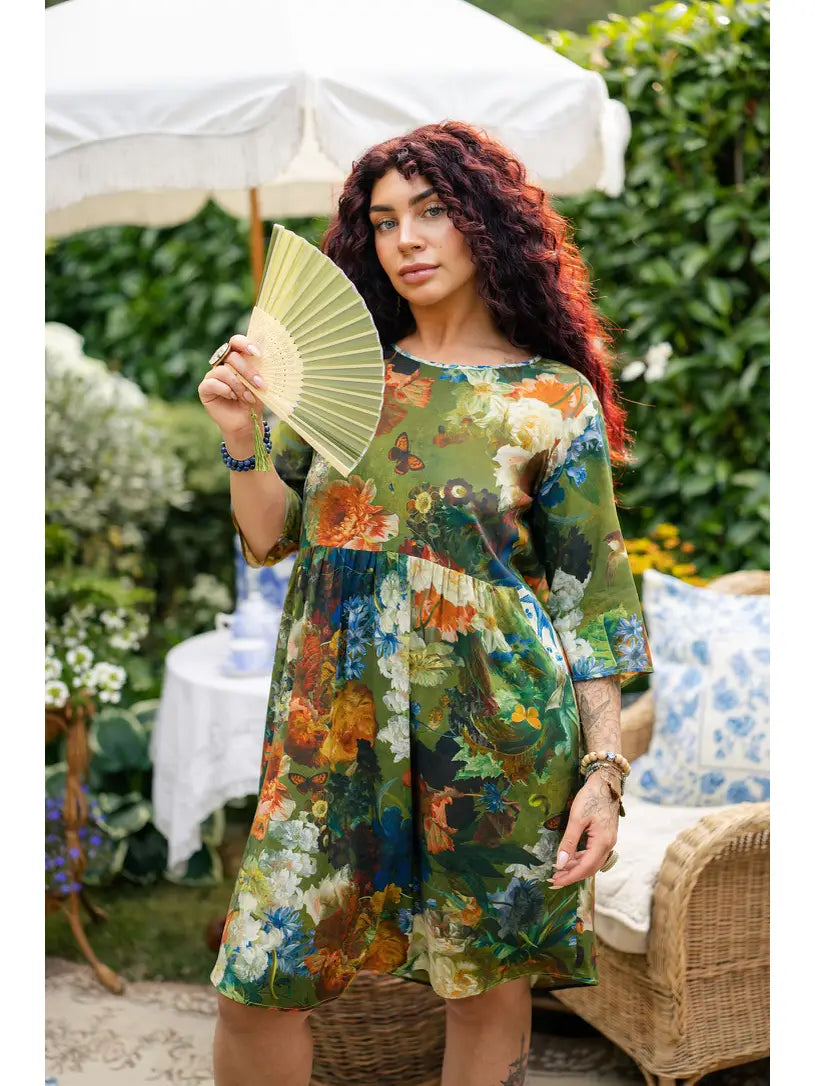 Curly-haired woman wearing a green floral artist dress tunic and holding a folding fan on a garden patio | Bliss by Cori