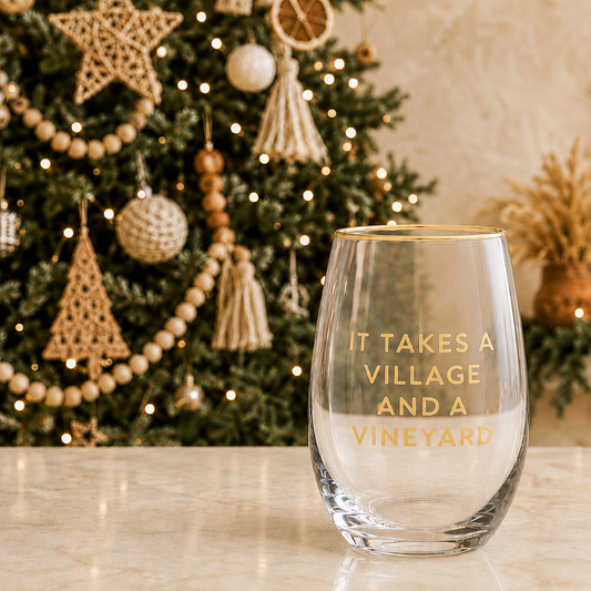 Clear stemless wine glass with gold rim and gold foil It Takes a Village and a Vineyard block text on marble counter in front of a boho Christmas tree with wood bead garland macrame tassels and warm fairy lights – Bliss by Cori
