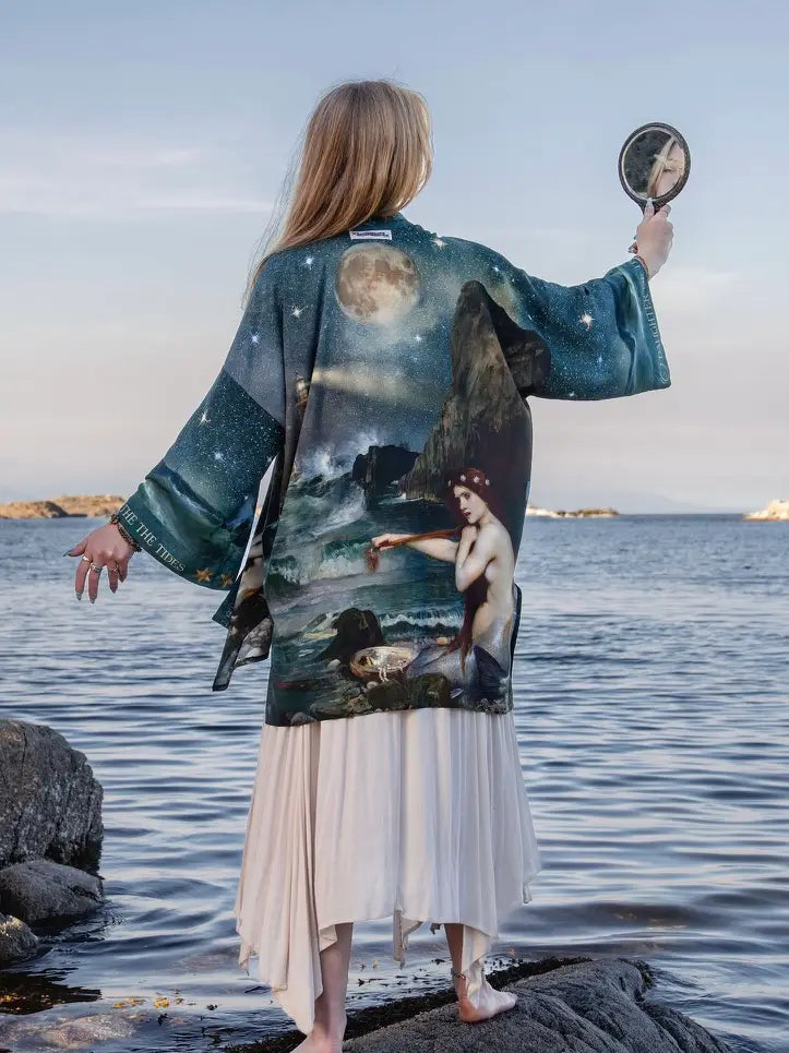 A woman stands on a rocky shoreline facing the sea, wearing a long-sleeved kimono-style cardigan with a detailed celestial and mermaid print. She holds a hand mirror in her raised hand to inspect the print on the back of the garment | Bliss by Cori