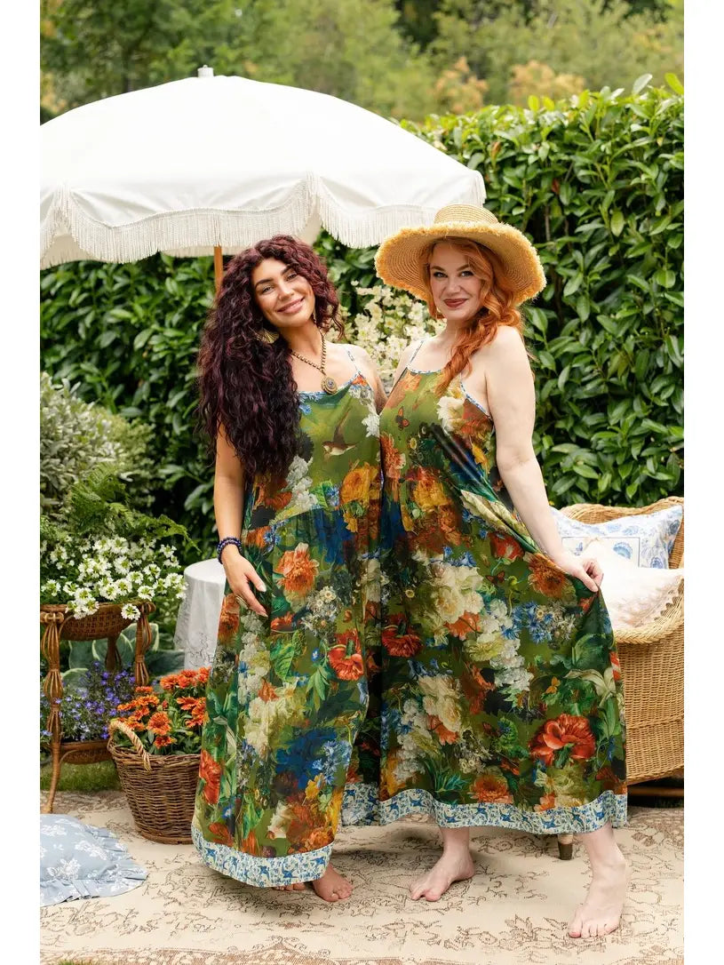 Two women modeling matching green floral-and-bird maxi slip dresses in a garden, with one wearing a wide-brimmed straw hat | Bliss by Cori