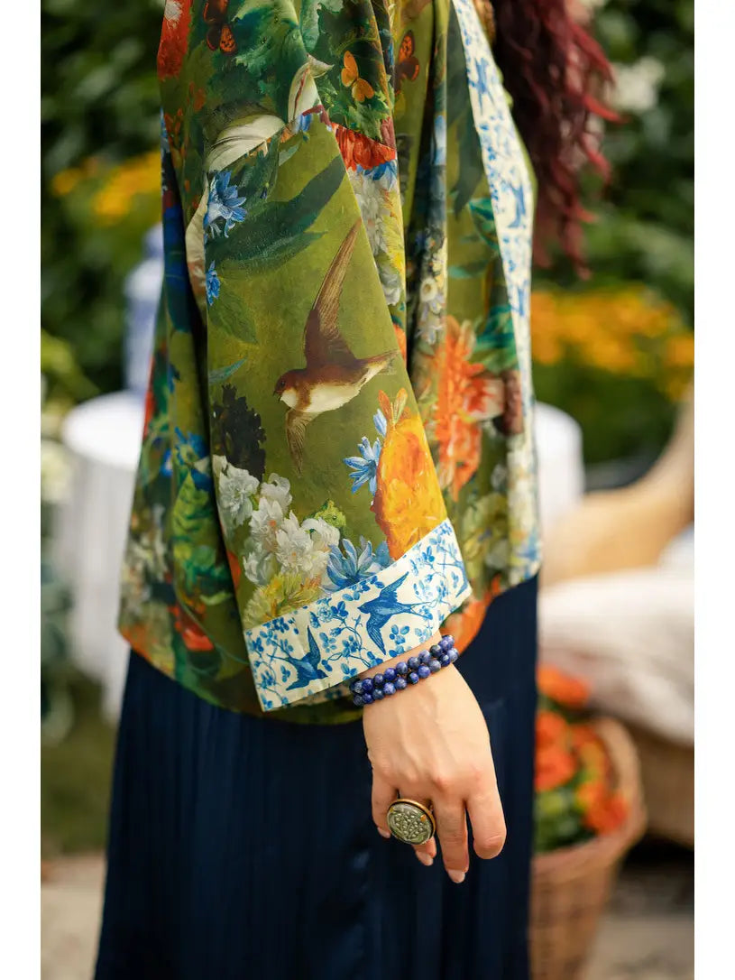 A close-up of a person wearing a green tropical print jacket with birds and flowers, featuring a blue and white patterned cuff, paired with a dark navy skirt | Bliss by Cori