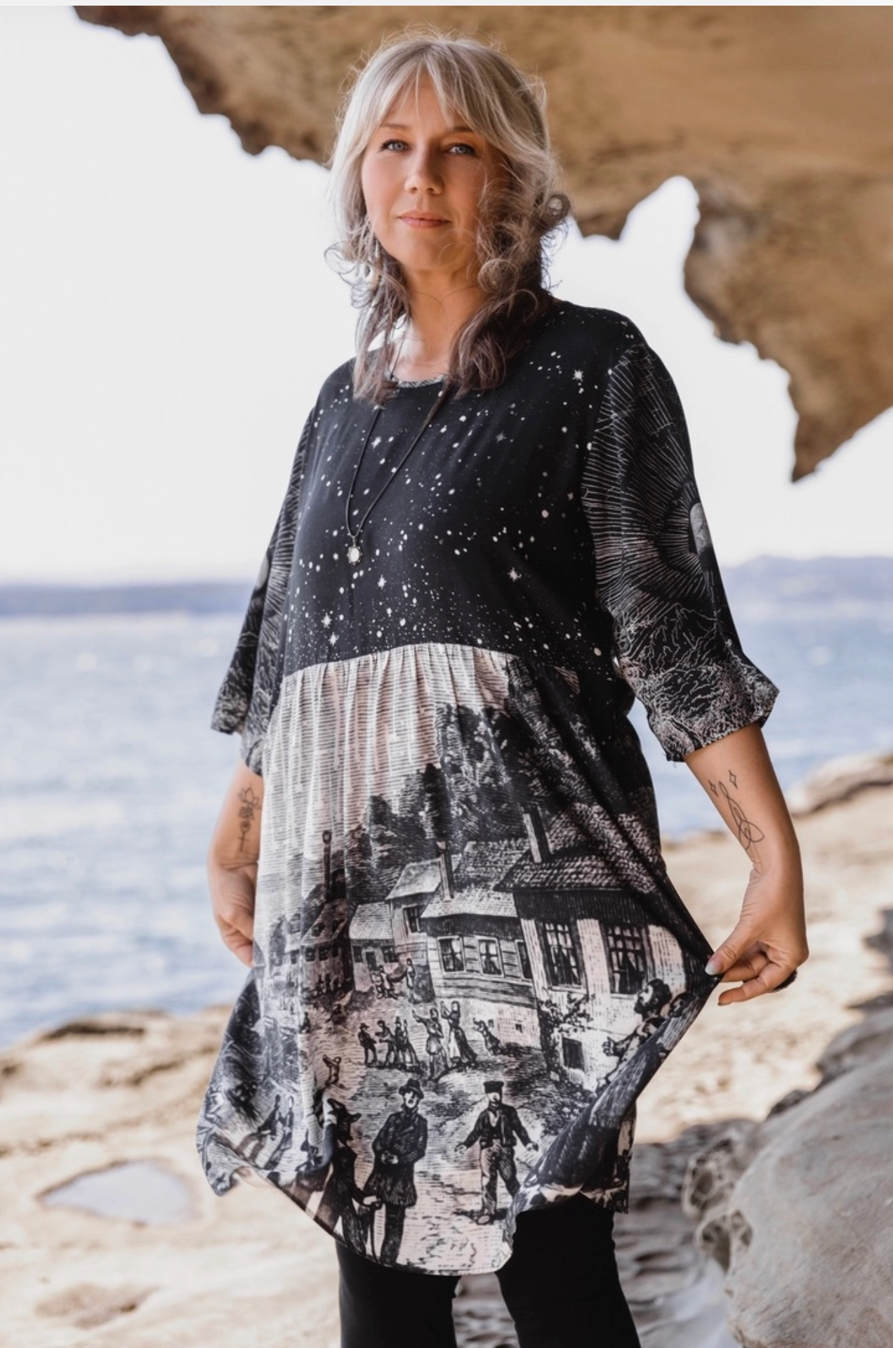 Front view of Stargazer Artist Dress on model standing on coastal rocks — star field bodice with Leonid meteor shower village scene skirt | Bliss by Cori Boutique