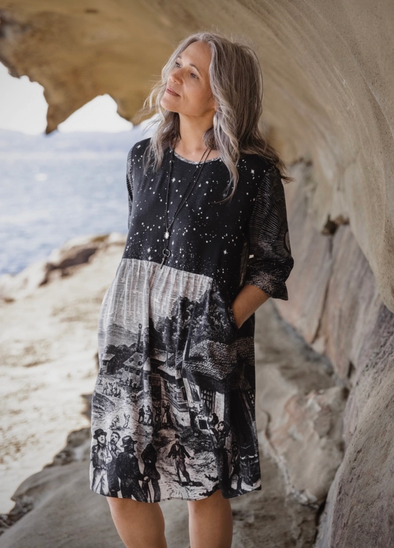 ide view of Stargazer Artist Dress with celestial star field bodice and village scene skirt, woman standing on coastal rocks | Bliss by Cori Boutique