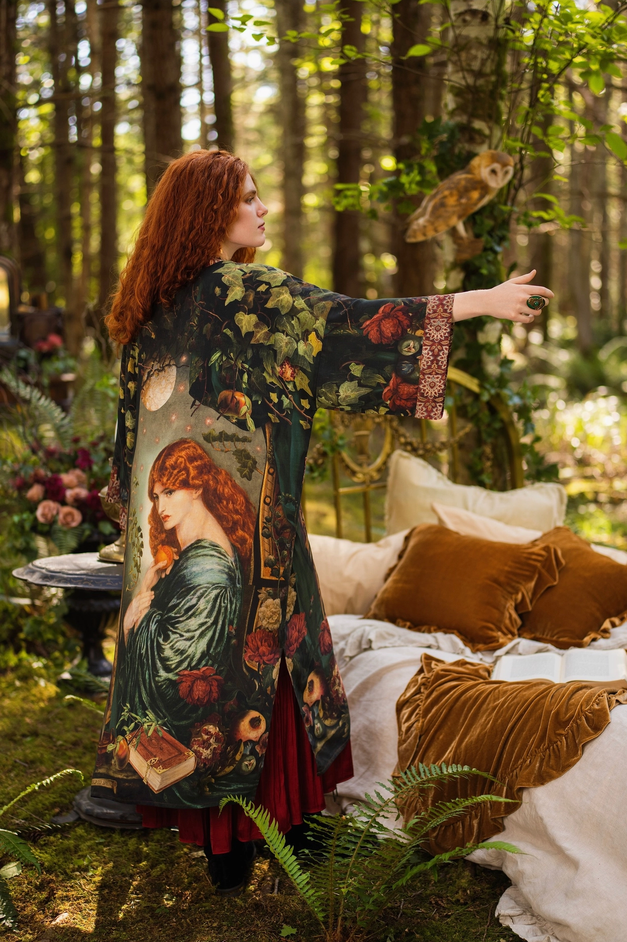 Back view of red-haired model in forest wearing Solstice Opera Duster Kimono showing full Rossetti Proserpine print, arm outstretched toward perched barn owl, velvet cushions in background — Bliss by Cori