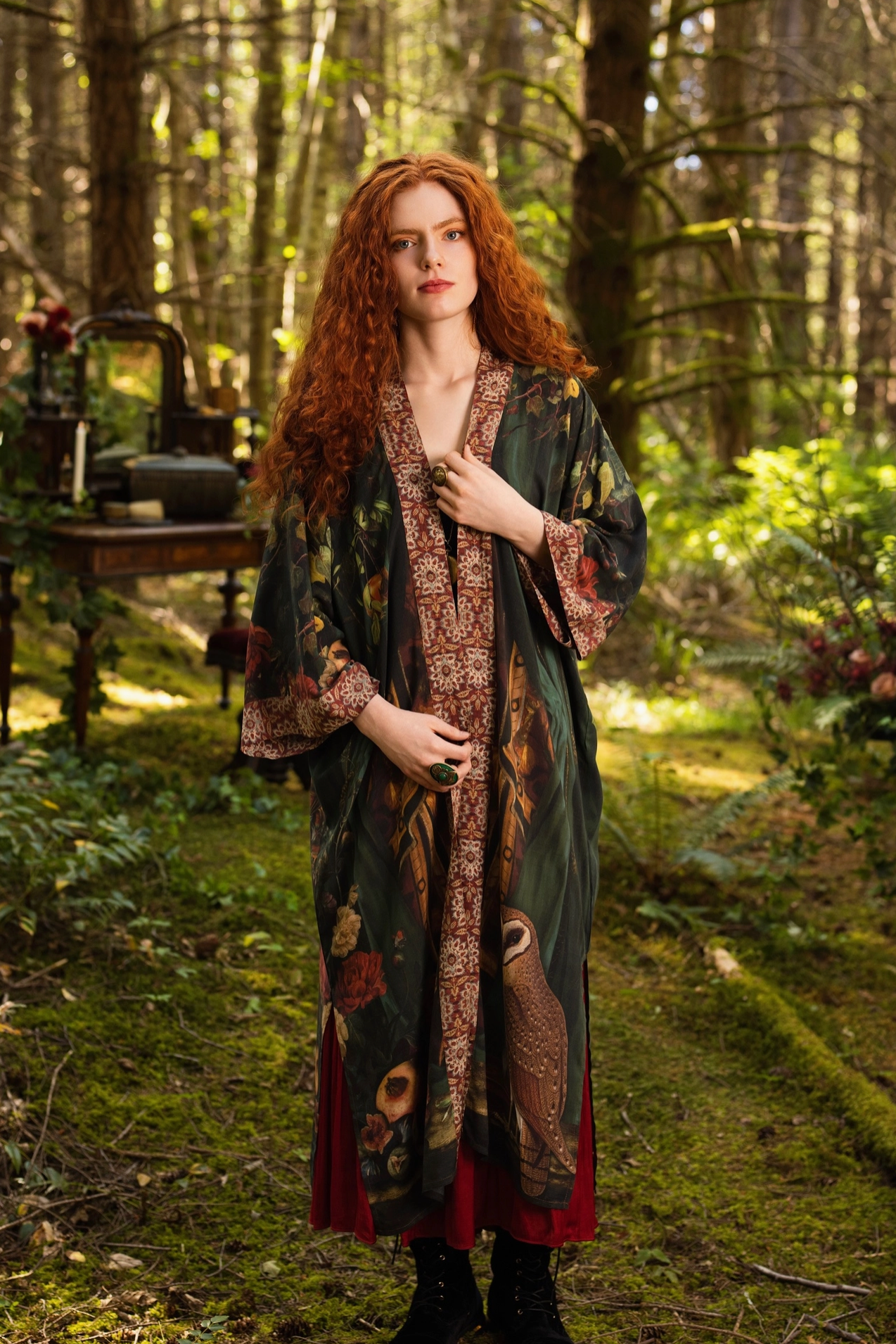 Red-haired model in sunlit forest wearing Solstice Opera Duster Kimono open over burgundy dress, owl and botanical print visible on front, antique writing desk in background — Bliss by Cori