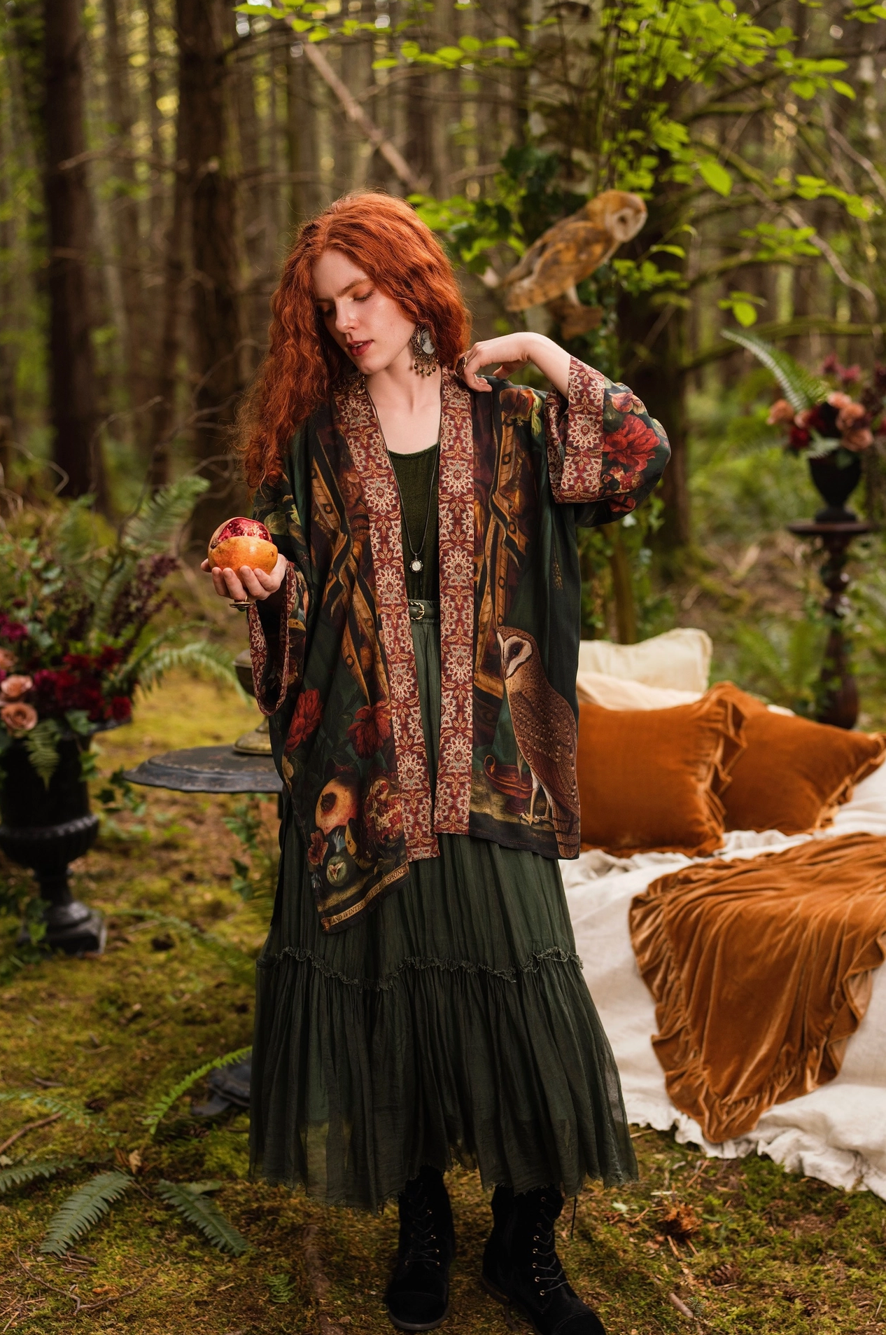 Front view of red-haired model in forest standing in Solstice Matinée Mid-Length Duster Kimono open over dark green dress, holding pomegranate, barn owl in background, velvet cushions and enchanted outdoor bedroom setting — Bliss by Cori