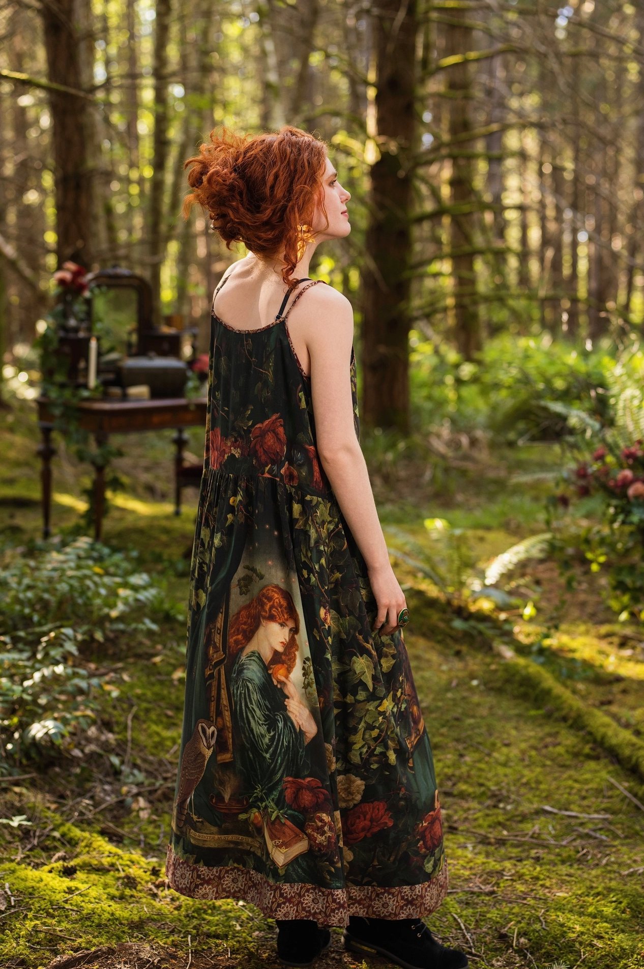 Side profile of red-haired model with upswept hair in sunlit forest wearing Solstice Bohéme Maxi Slip Dress, Rossetti Proserpine portrait print cascading to tapestry hem, antique writing desk with candle behind — Bliss by Cori
