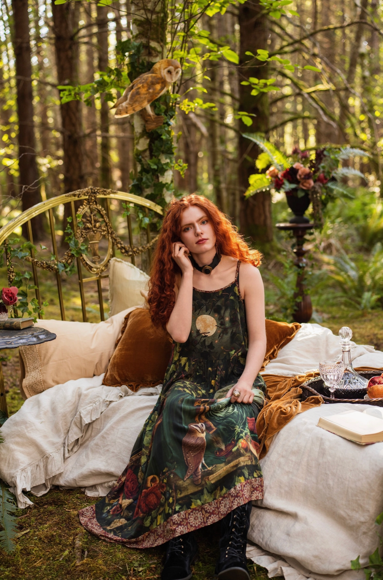 Red-haired model seated on antique brass bed in forest wearing Solstice Bohéme Maxi Slip Dress, barn owl perched in tree above, pomegranate and roses on tray, velvet cushions and open book nearby — Bliss by Cori