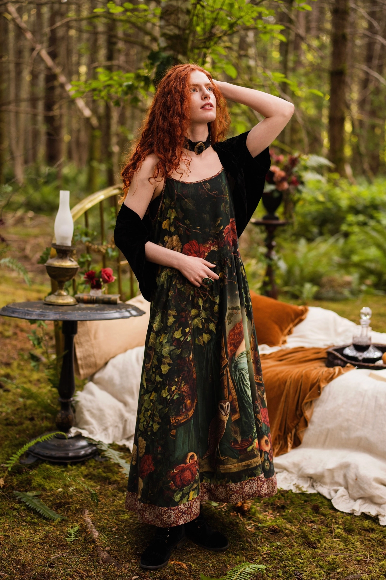 Full-length view of red-haired model in forest wearing Solstice Bohéme Maxi Slip Dress layered under a black kimono, Proserpine print and tapestry hem visible, oil lamp and outdoor bed behind — Bliss by Cori