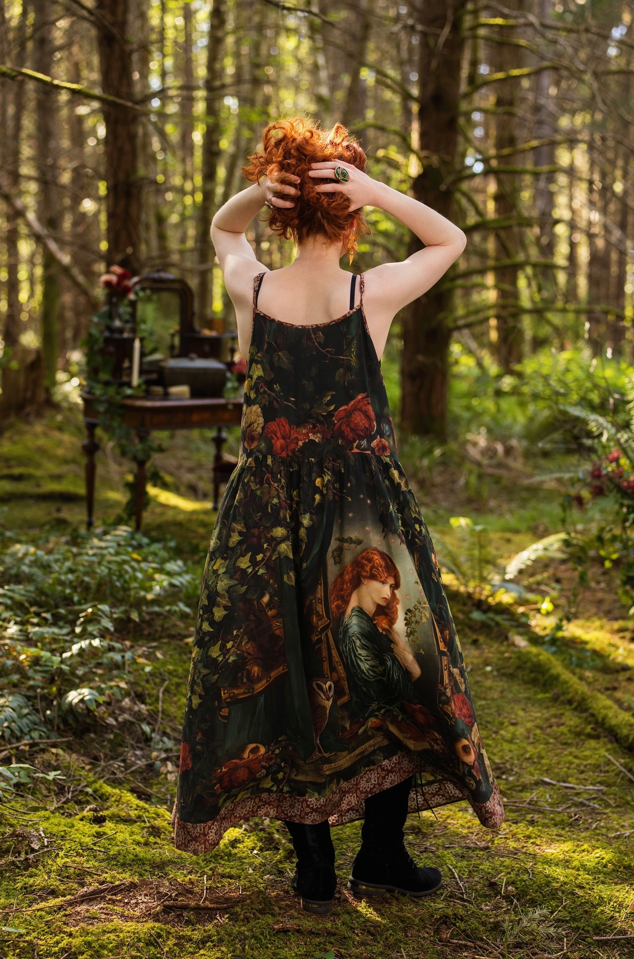 Three-quarter view of red-haired model in forest wearing Solstice Bohéme Maxi Slip Dress, showing Rossetti Proserpine print and tapestry hem, enchanted outdoor bedroom with roses and velvet cushions — Bliss by Cori