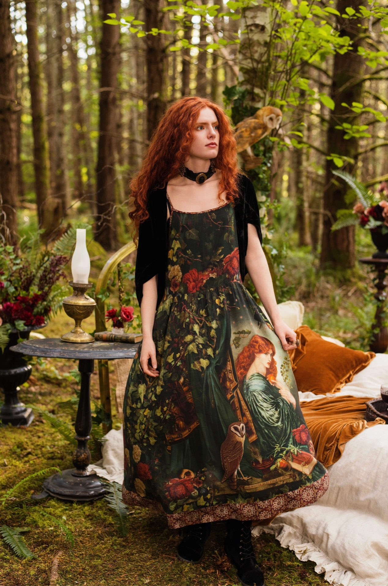 Full-length front view of red-haired model in forest wearing Solstice Bohéme Maxi Slip Dress, showing full moon, ivy and Proserpine print with tapestry border hem, oil lamp and antique bed behind — Bliss by Cori