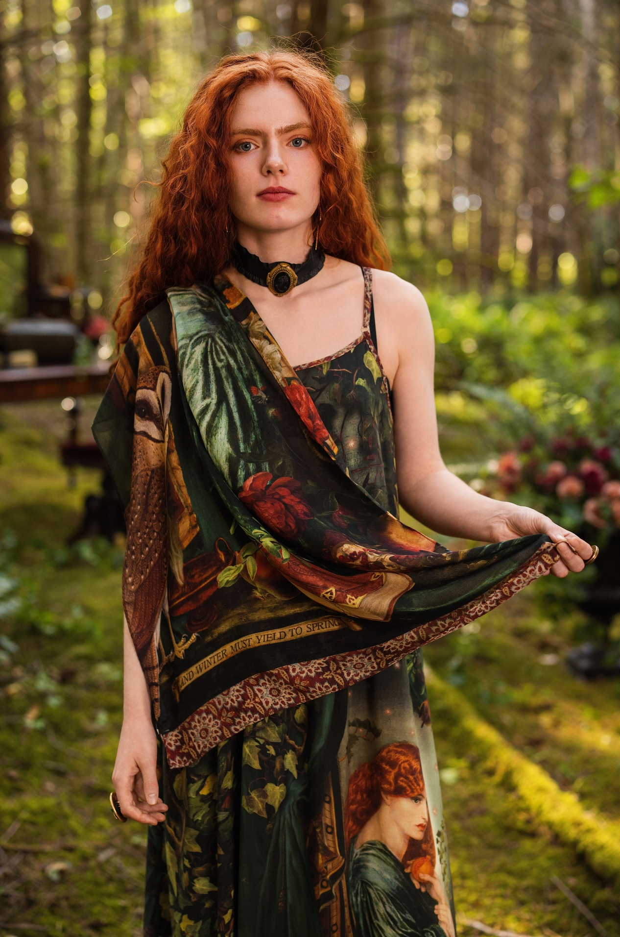 Red-haired model in sunlit forest wearing Solstice Bohemian Scarf draped over one shoulder over Solstice Maxi Slip Dress, Rossetti Proserpine print and "and winter must yield to spring" banner visible, enchanted garden behind — Bliss by Cori