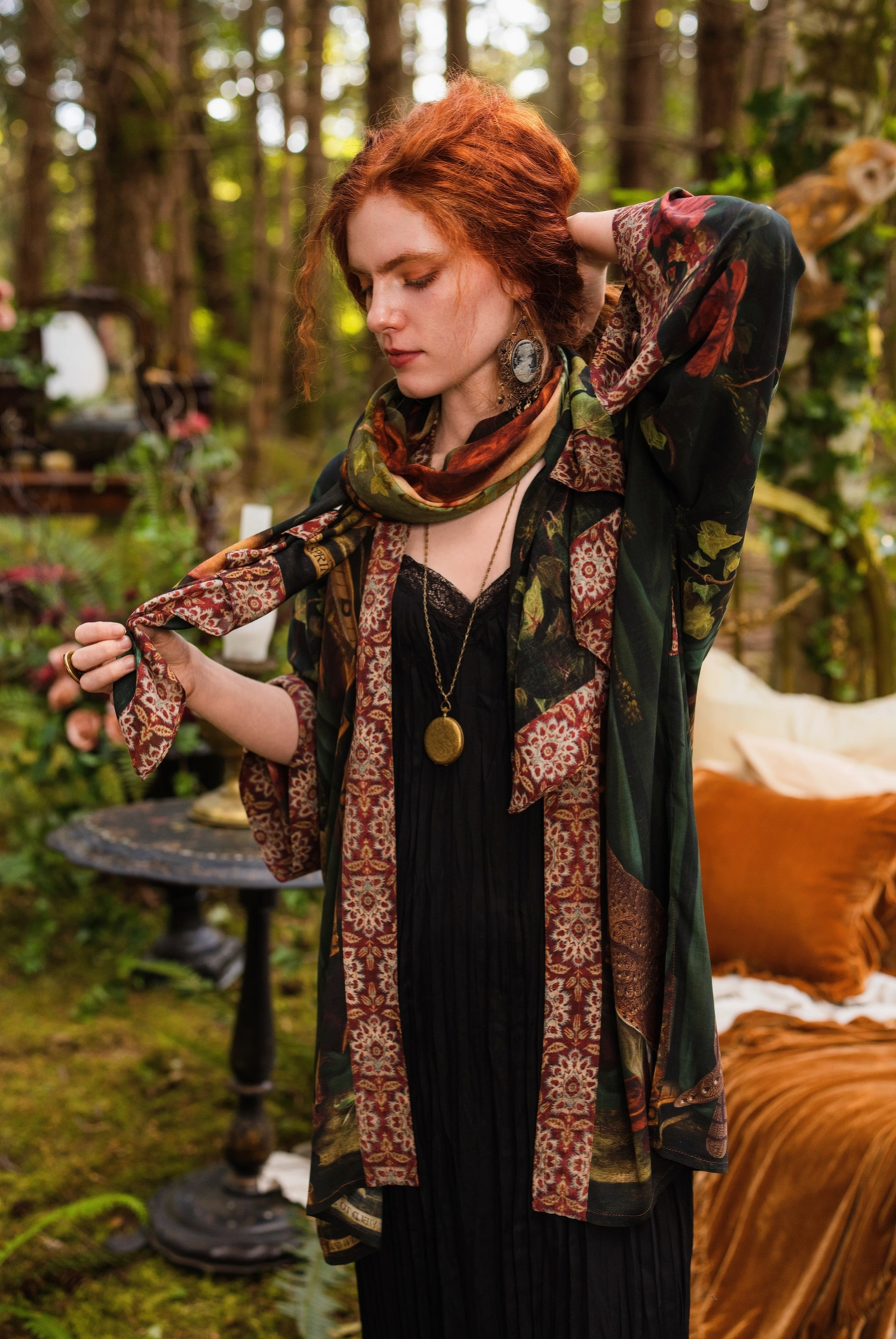 Red-haired model with upswept hair in forest garden styling Solstice Bohemian Scarf loosely around neck over Solstice Kimono and black maxi dress, tapestry border and Persephone print visible, enchanted outdoor bedroom setting behind — Bliss by Cori