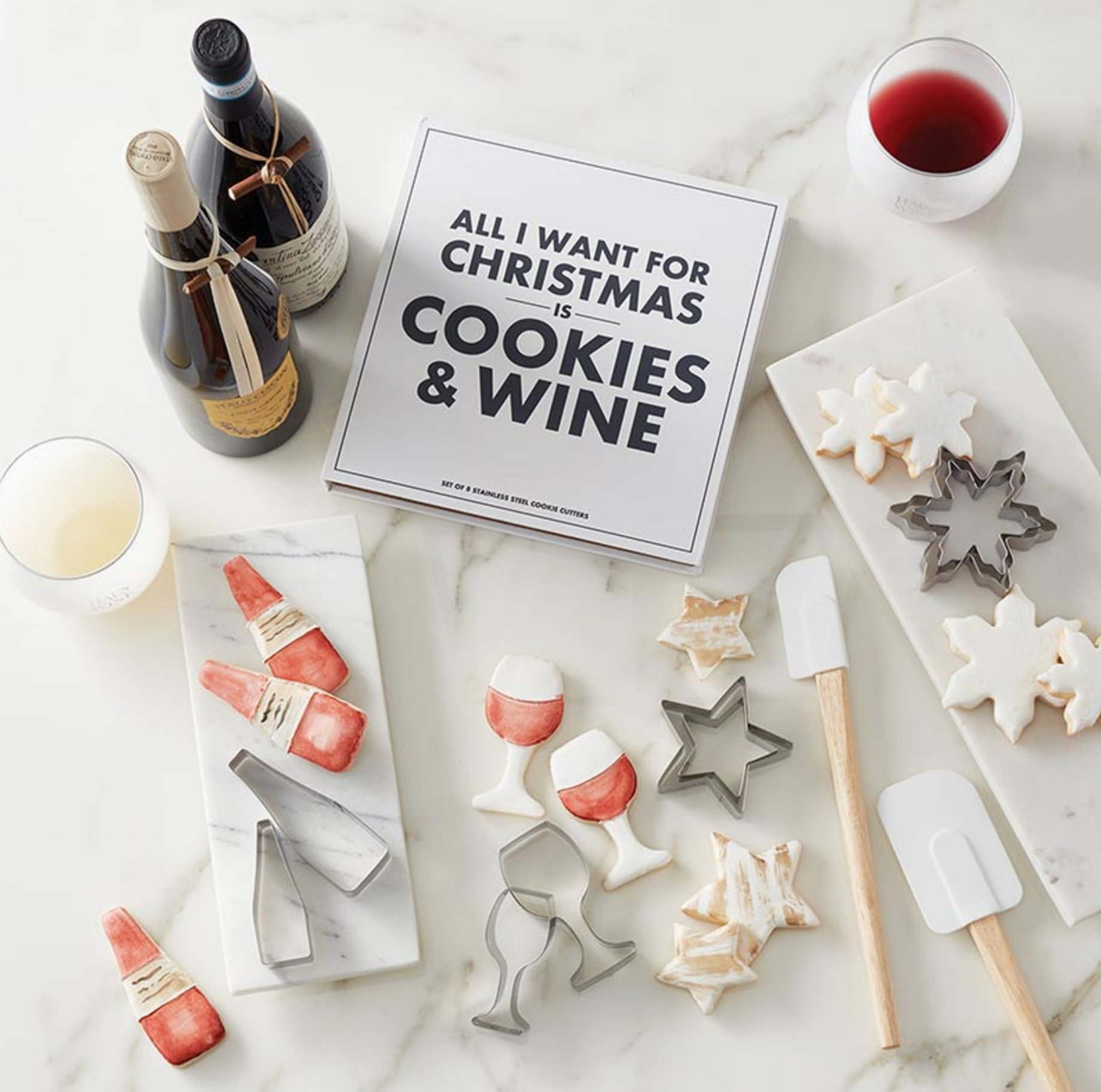 Overhead flat lay of All I Want for Christmas is Cookies and Wine book box gift set surrounded by wine bottles glasses iced sugar cookies shaped as wine glasses and bottles and stainless steel cookie cutters on marble – Bliss by Cori
