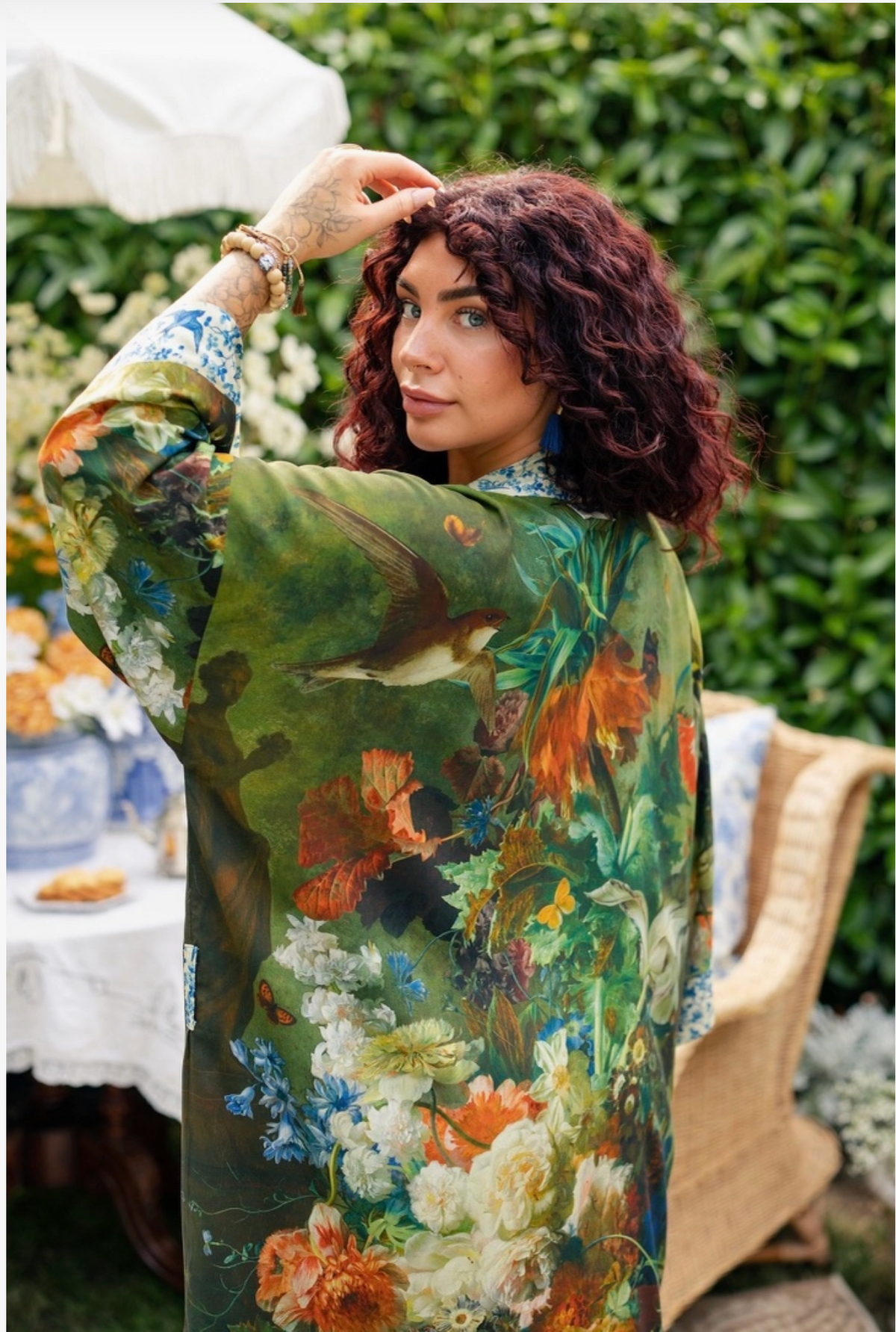 A woman wearing a long-sleeve kaftan-style dress with a vibrant, detailed botanical and bird print, photographed in an outdoor garden setting | Bliss by Cori
