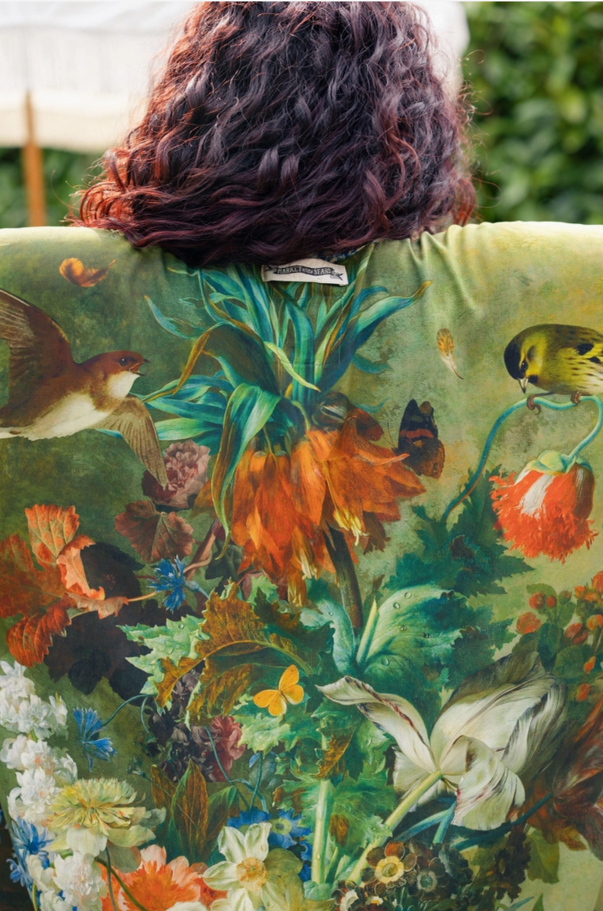 A person viewed from behind wearing a green printed chair or cushion with a detailed botanical and avian design featuring flowers, leaves, butterflies, and birds | Bliss by Cori