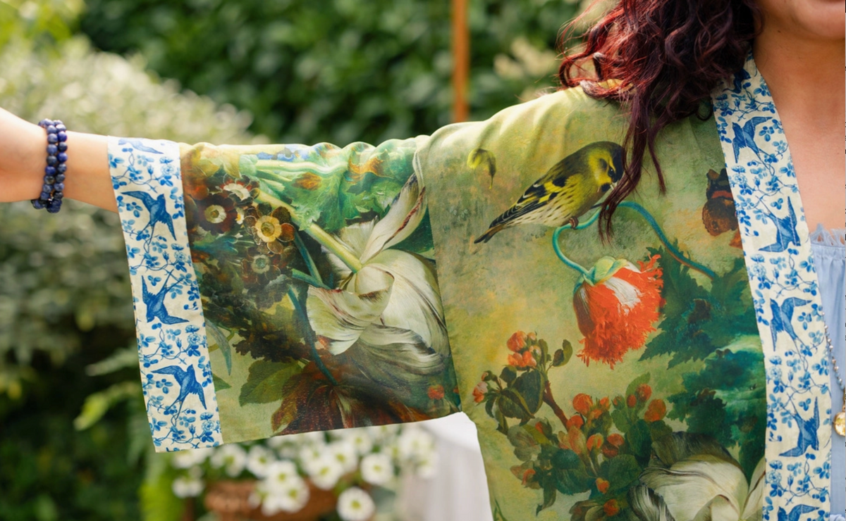 A close-up of a person wearing a lightweight open-front kimono-style jacket with wide sleeves. The garment features a detailed vintage-style botanical and bird print in green, cream, red, and blue, with blue-and-white patterned cuffs and trim | Bliss by Cori