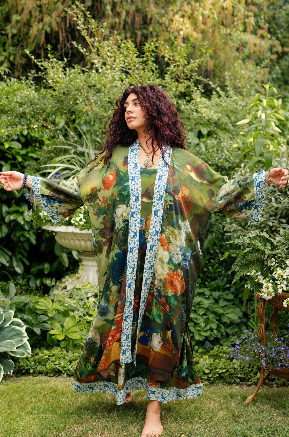 A woman stands barefoot in a lush garden wearing a long, flowing floral robe with blue patterned trim. The robe features a multicolor print with green, orange, and red tones | Bliss by Cori