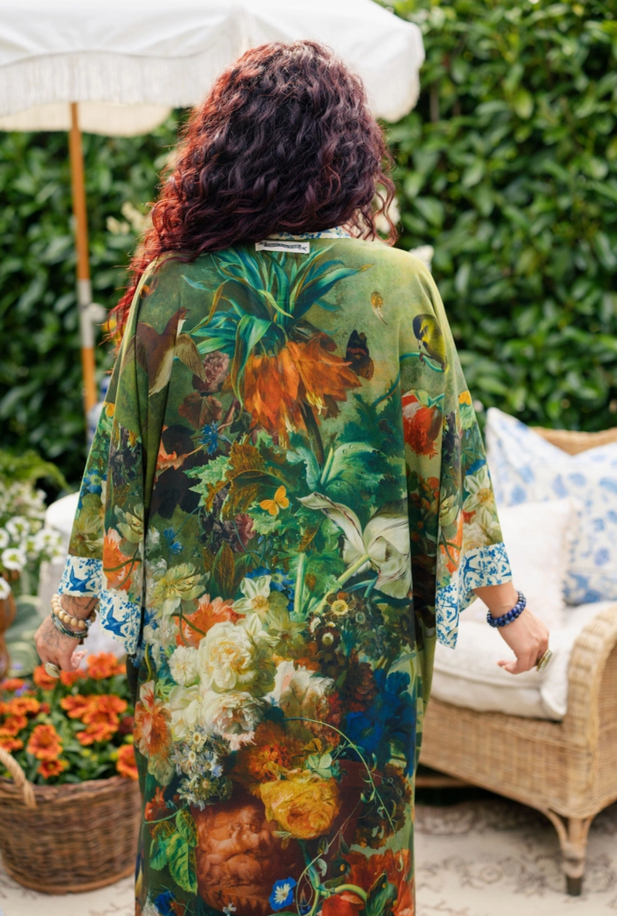 A woman viewed from behind wearing a flowing kaftan-style dress with a dense floral and botanical print in green, orange, cream, blue, and brown tones. She is standing outdoors on a patio with a wicker chair, flowers, and greenery in the background | Bliss by Cori