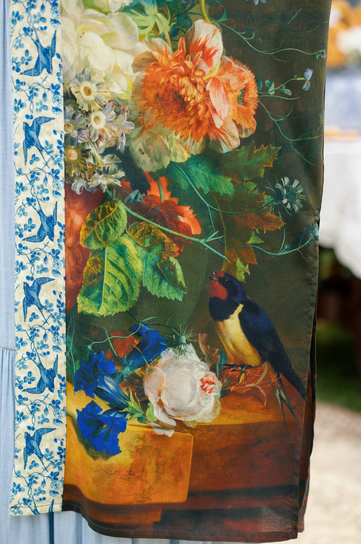 A close-up of a printed textile featuring a classical-style floral still life with a bird, alongside a blue-and-white patterned border. The visible design includes large orange and white flowers, green leaves, blue blossoms, and a dark background with a bird in blue, yellow, and red tones | Bliss by Cori