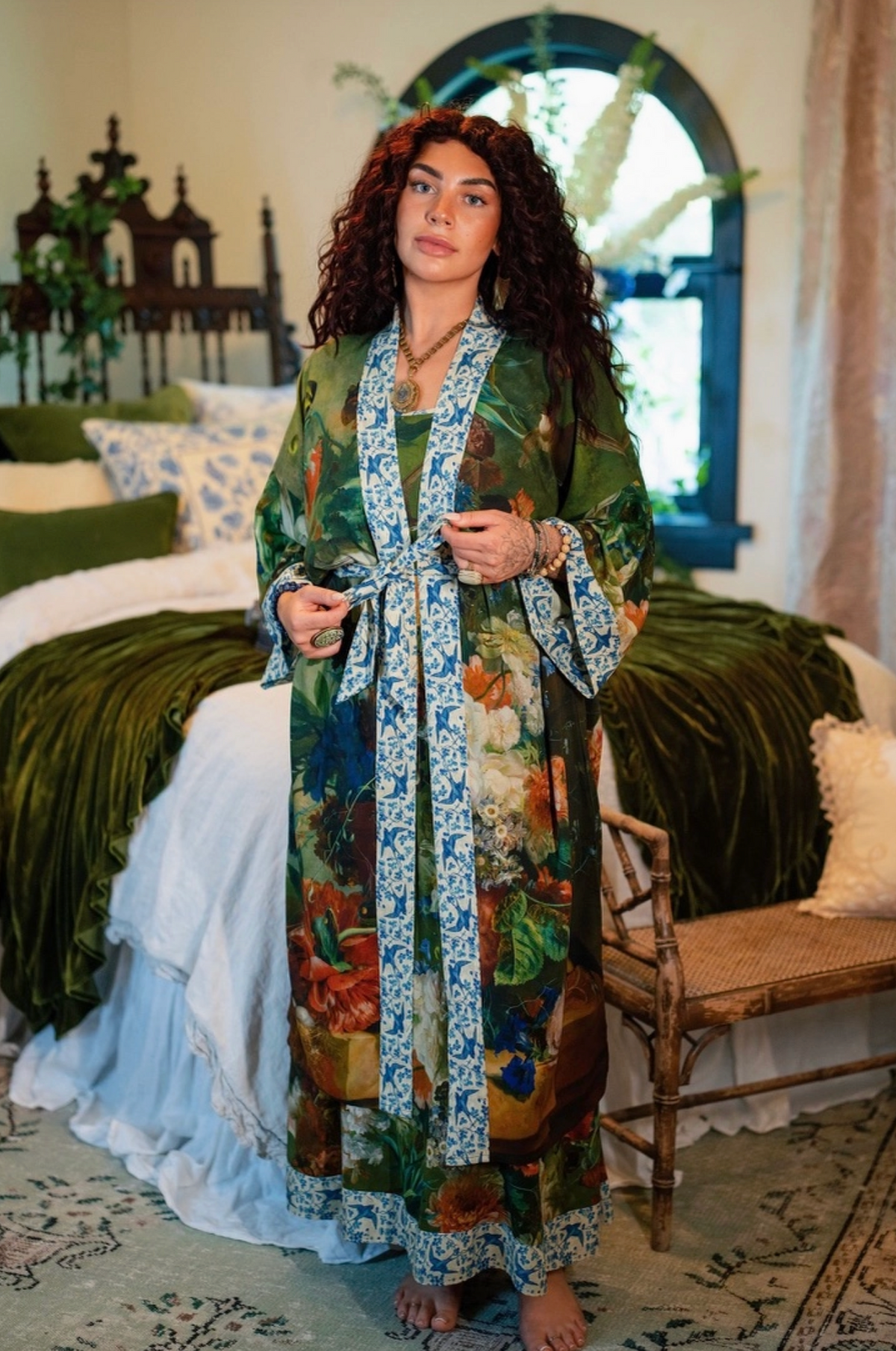 A woman stands in a bedroom wearing a long, colorful floral robe with blue patterned trim. The robe features a mix of green, orange, red, and blue botanical prints with blue patterned borders | Bliss by Cori