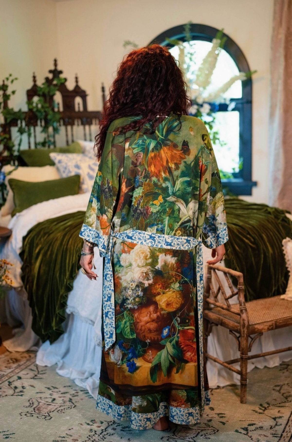 A woman viewed from behind wearing a long, flowing floral robe in a decorated bedroom. The robe features a rich botanical print with green, orange, yellow, and blue tones, and is styled with a blue-and-white patterned belt and matching patterned hems | Bliss by Cori