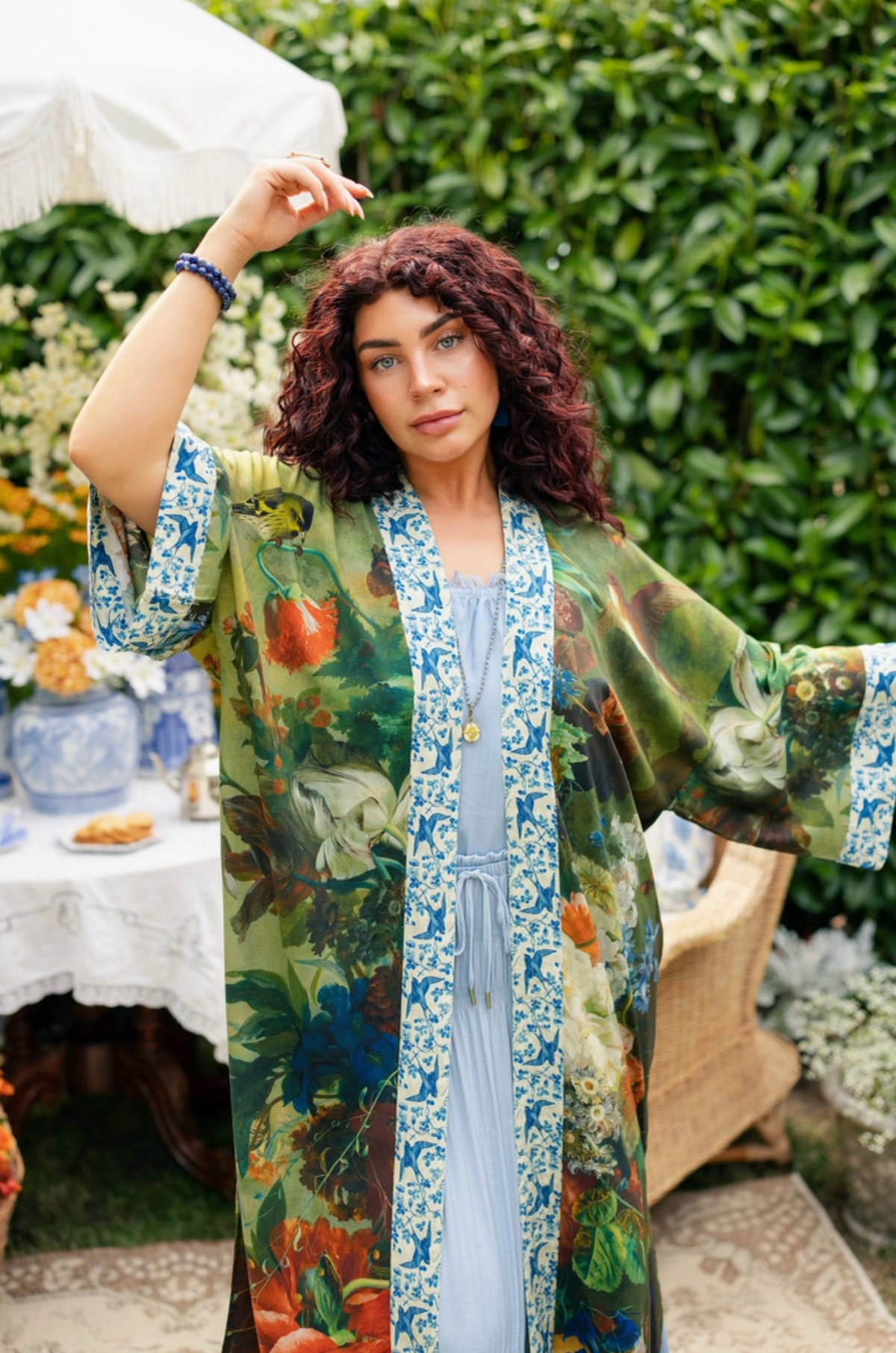 A woman stands outdoors in a garden setting wearing a long, open-front kaftan robe with a colorful botanical print over a light blue dress. The robe features blue patterned trim and a drawstring tie at the front | Bliss by Cori