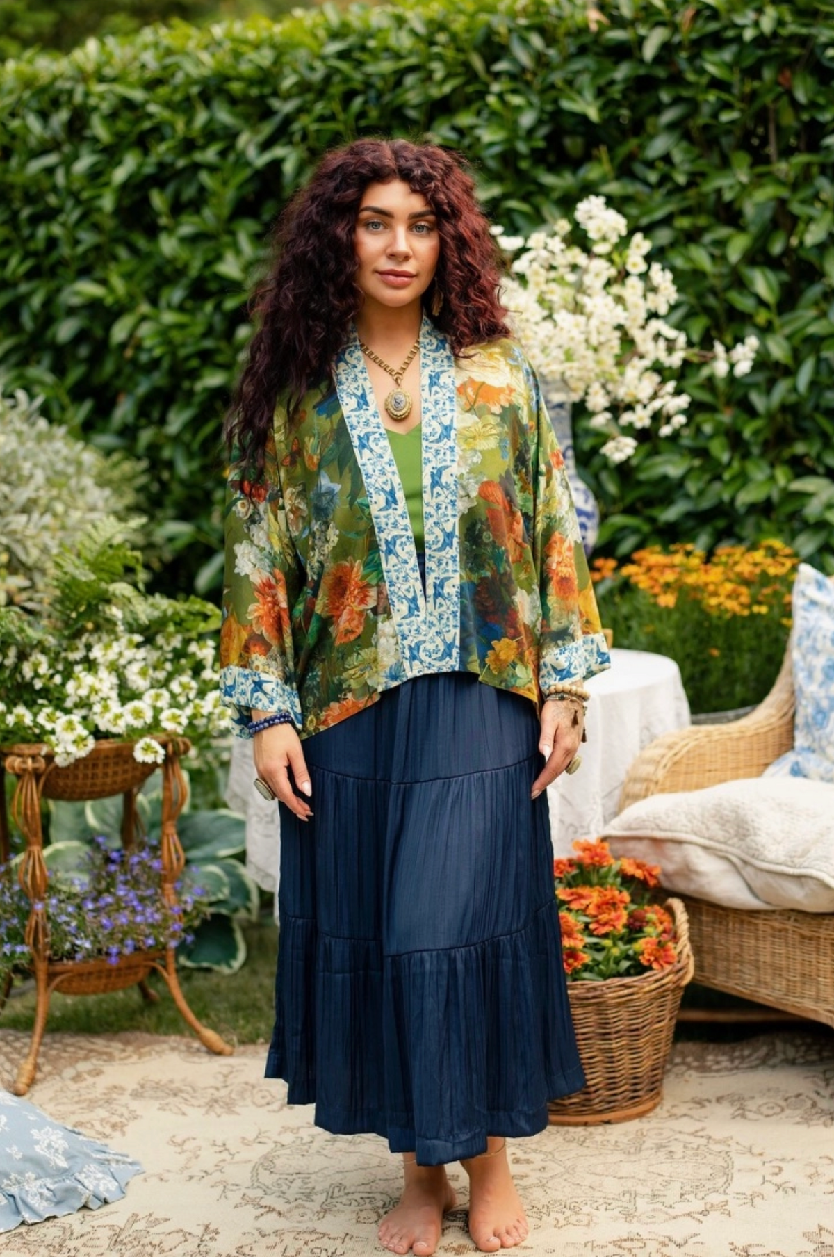 A woman stands barefoot in a garden courtyard wearing a bohemian-style outfit consisting of a sheer multicolor floral print jacket over a green top and a long navy blue pleated skirt | Bliss by Cori
