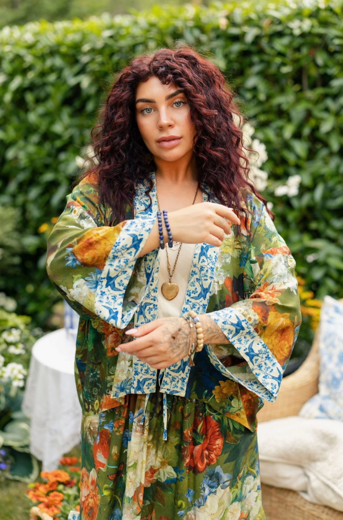 A woman with curly reddish-brown hair stands outdoors in a garden wearing a bohemian-style floral robe. The robe features a green base with blue, orange, and yellow patterns, layered over a white top | Bliss by Cori